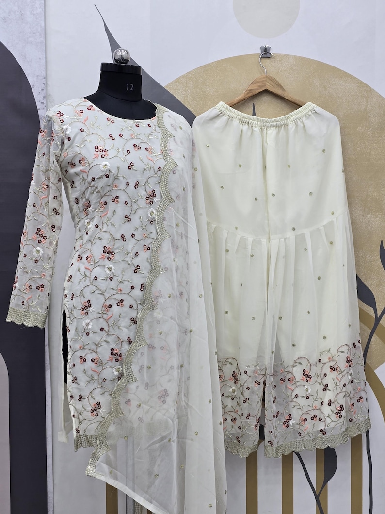 May include: A white embroidered salwar kameez set. The long-sleeved tunic and matching pants are adorned with floral embroidery and sequins. The set includes a sheer dupatta with a scalloped edge.