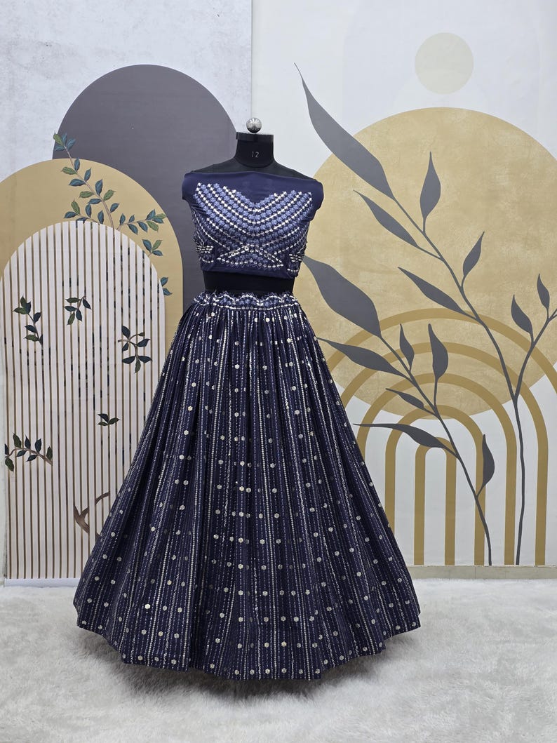 May include: A navy blue two-piece formal outfit. The top is a cropped, sleeveless design with silver embellishments. The skirt is a full, floor-length style with vertical stripes and scattered silver dots. The outfit is displayed on a mannequin.