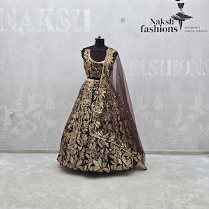 Peut inclure: Un lehenga marron et doré exposé sur un mannequin. Le lehenga présente des broderies dorées complexes en forme de feuilles. Un dupatta marron assorti avec une bordure dorée drape le mannequin. Le logo de Naksh Fashions est visible en arrière-plan.