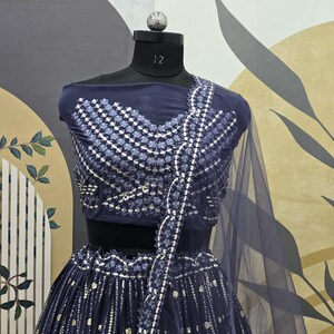 May include: A navy blue two-piece formal outfit displayed on a mannequin. The top features a geometric pattern with silver accents, and the skirt has vertical silver embellishments. A sheer, navy blue scarf drapes over the shoulder.