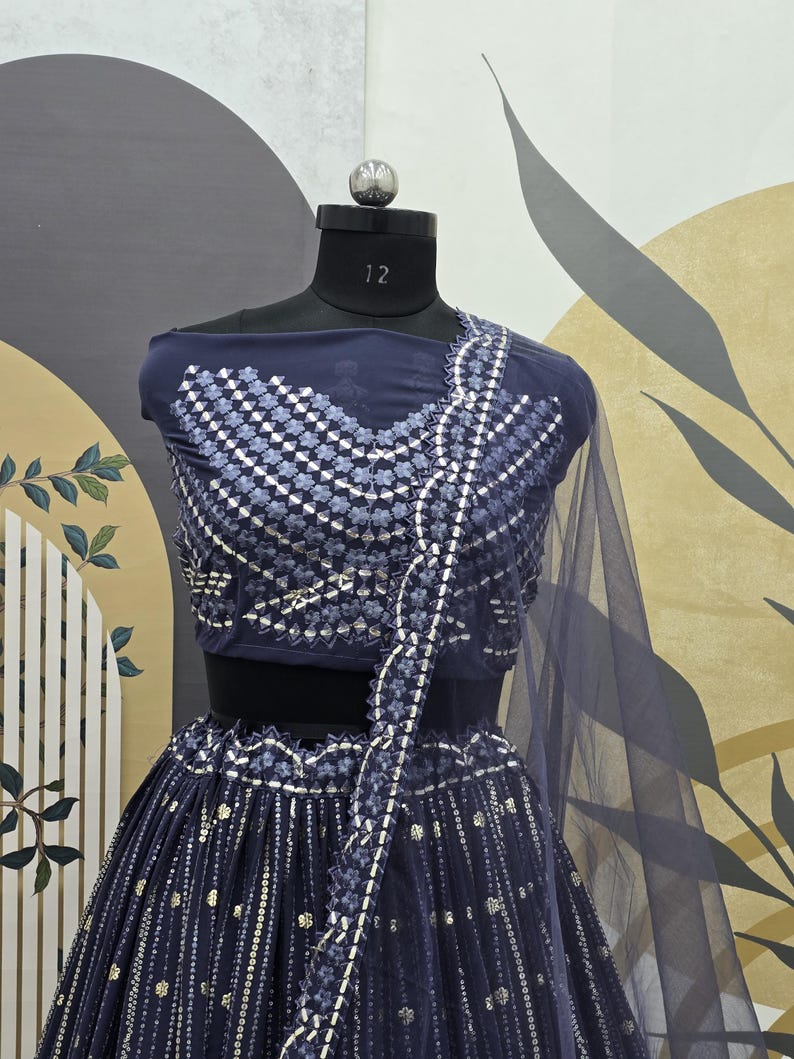 May include: A navy blue two-piece formal outfit displayed on a mannequin. The top features a geometric pattern with silver accents, and the skirt has vertical silver embellishments. A sheer, navy blue scarf drapes over the shoulder.