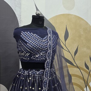 May include: A navy blue lehenga choli set displayed on a mannequin. The top features intricate silver embroidery, and the skirt has vertical silver embellishments. A sheer navy blue dupatta is draped over the shoulder. The mannequin is set against a neutral background.
