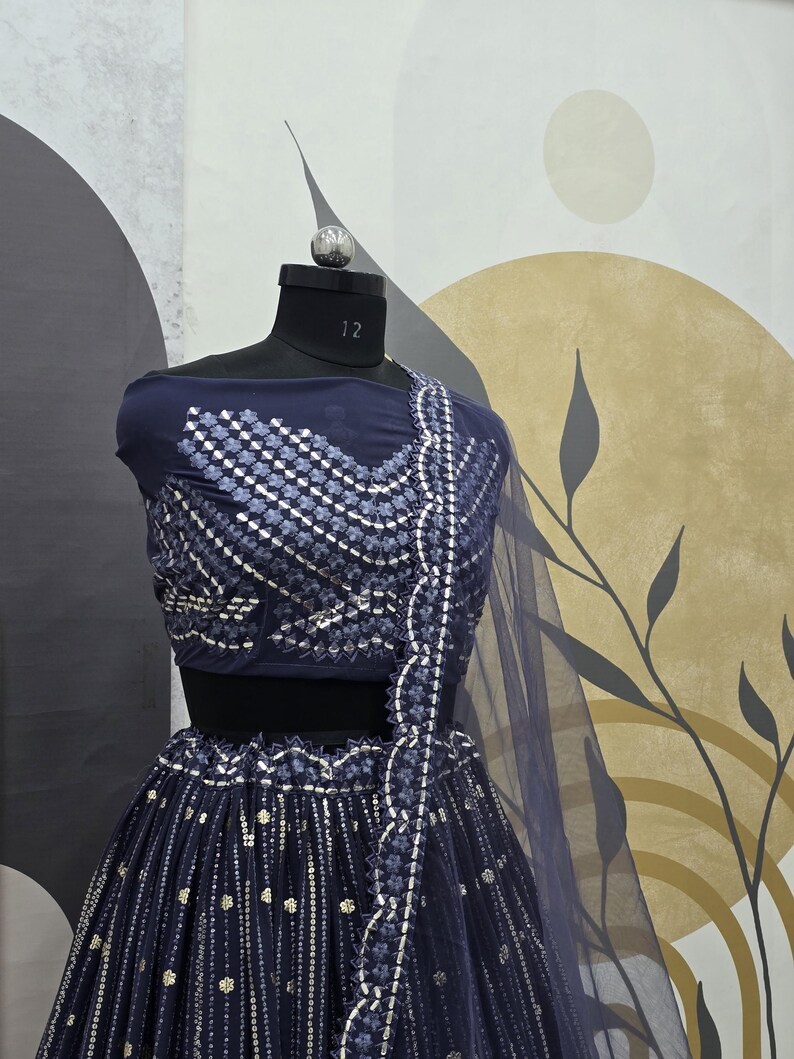 May include: A navy blue lehenga choli set displayed on a mannequin. The top features intricate silver embroidery, and the skirt has vertical silver embellishments. A sheer navy blue dupatta is draped over the shoulder. The mannequin is set against a neutral background.