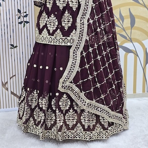 May include: A maroon-colored dress with gold embroidery and a matching dupatta. The dress has a flared skirt and a fitted bodice. The dupatta is sheer and has a delicate border.