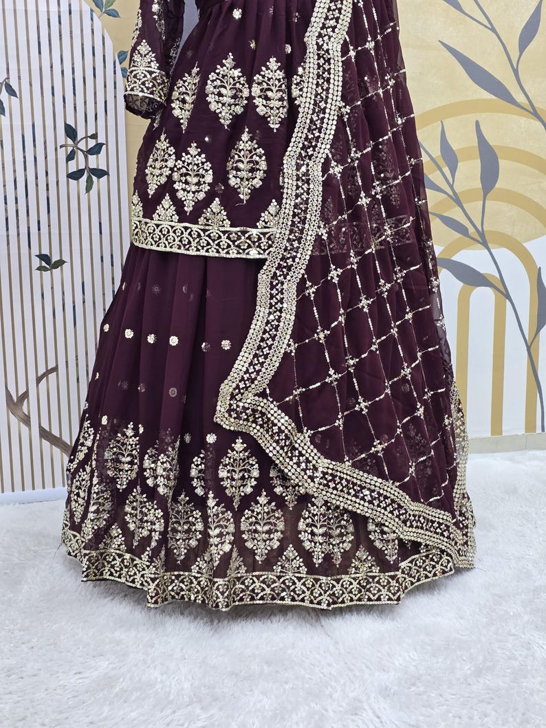 May include: A maroon-colored dress with gold embroidery and a matching dupatta. The dress has a flared skirt and a fitted bodice. The dupatta is sheer and has a delicate border.