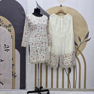 May include: A cream-colored, long-sleeved tunic and matching palazzo pants set. The tunic is adorned with floral embroidery and a scalloped hem. The palazzo pants have a similar embroidered design along the hemline. The outfit is displayed on a mannequin and hanger.