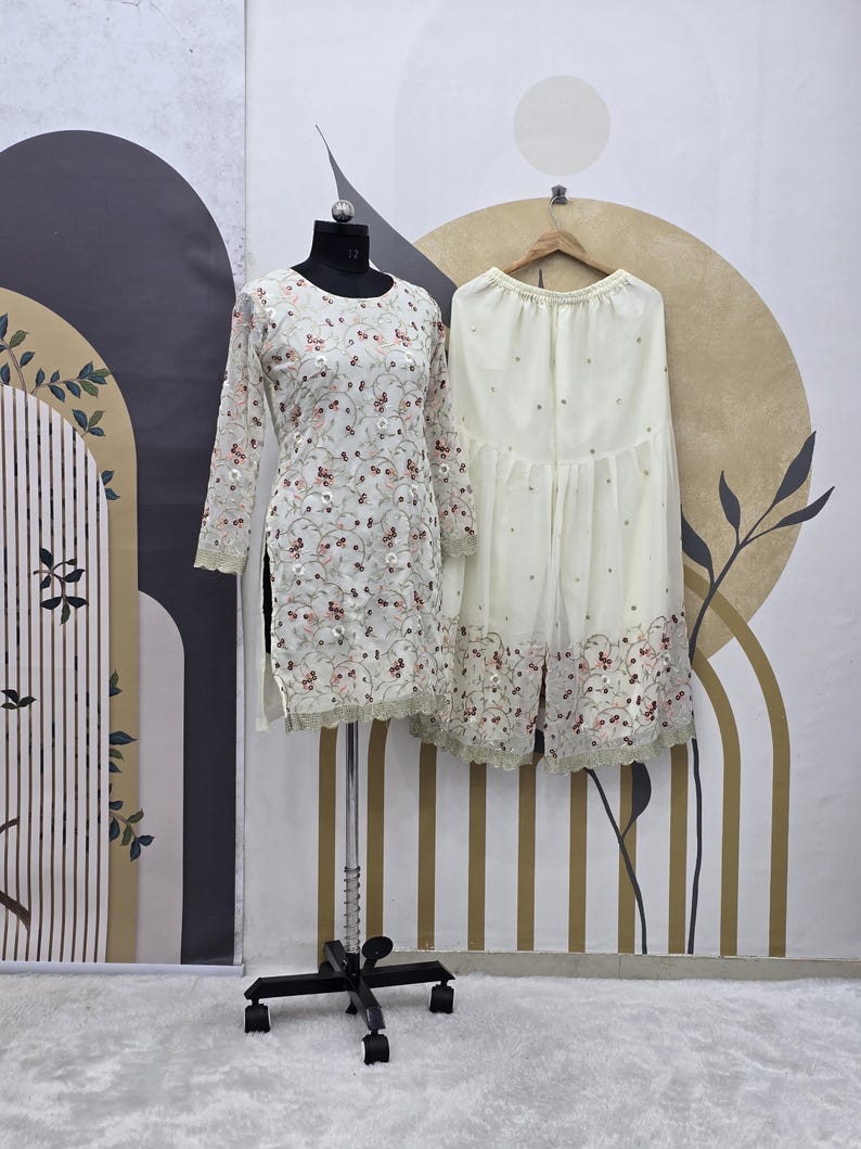 May include: A cream-colored, long-sleeved tunic and matching palazzo pants set. The tunic is adorned with floral embroidery and a scalloped hem. The palazzo pants have a similar embroidered design along the hemline. The outfit is displayed on a mannequin and hanger.
