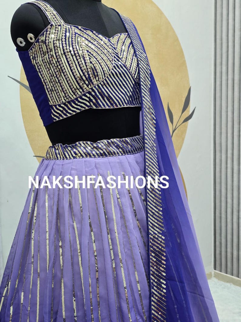Op de afbeelding: Een paars en blauwe set met een crop top en rok met een bijpassende dupatta. De top heeft een V-hals en de rok is een vloeiende A-lijn stijl. De set is versierd met zilveren pailletten.