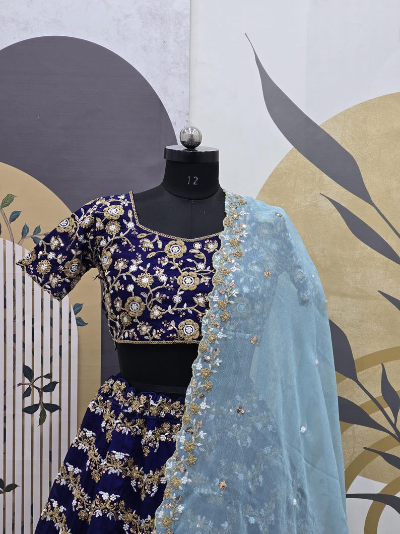 May include: A dark blue embroidered lehenga with a matching choli and a light blue dupatta. The choli has a floral design with gold and white embellishments. The lehenga and dupatta are also embroidered with gold and white thread.