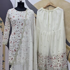 May include: A cream-colored, embroidered salwar kameez set. The long-sleeved top and dupatta feature floral embroidery with pink and maroon accents. The matching palazzo pants are also cream-colored and have a similar embroidered border.