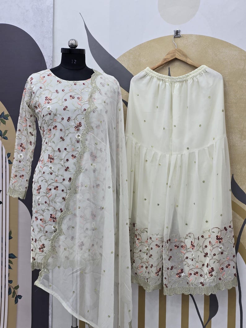 May include: A cream-colored, embroidered salwar kameez set. The long-sleeved top and dupatta feature floral embroidery with pink and maroon accents. The matching palazzo pants are also cream-colored and have a similar embroidered border.