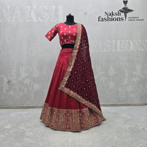 May include: A red lehenga choli set on a mannequin. The set includes a short-sleeved top, a long skirt, and a sheer maroon dupatta. The skirt features gold embroidery along the hem. The top has gold embroidery. The dupatta has gold embellishments.