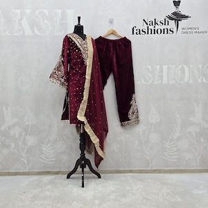 May include: A burgundy velvet salwar kameez set displayed on a mannequin. The set includes a tunic with gold embroidery, matching pants, and a sheer dupatta. The outfit is embellished with gold trim and beadwork. The logo "Naksh fashions" is visible in the background.