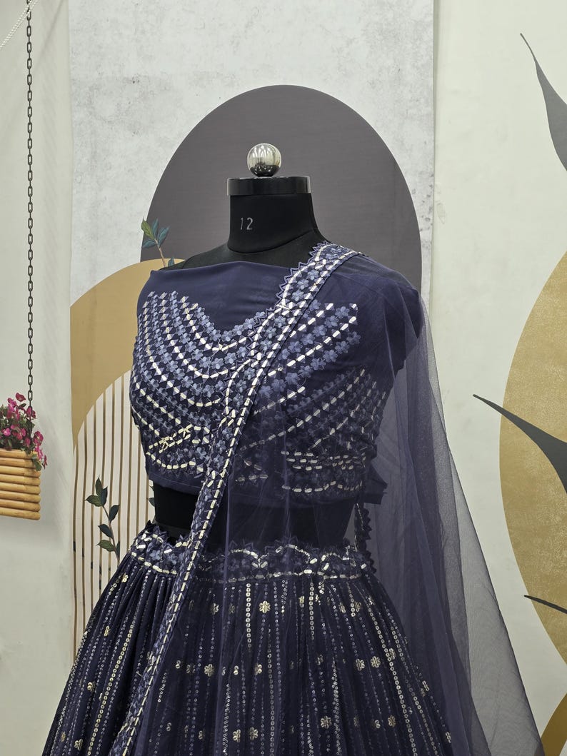 May include: A navy blue formal dress with intricate silver embroidery. The dress features a fitted bodice and a flowing skirt, with a matching sheer shawl. The ensemble is displayed on a mannequin, showcasing the detailed design and elegant style.