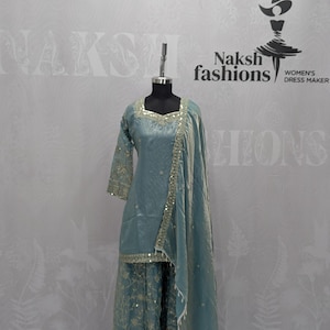 May include: A dusty blue three-piece outfit on a mannequin, featuring a square-neck tunic with gold embroidery and mirror work, wide-leg trousers with matching floral patterns, and a coordinating dupatta draped over one shoulder.