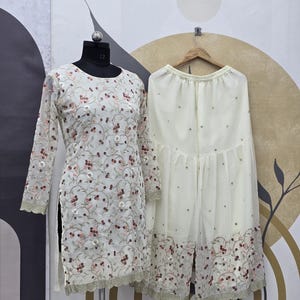 May include: A cream-colored embroidered tunic and matching palazzo pants set. The tunic features long sleeves and a floral pattern with pink accents. The palazzo pants have a similar embroidered border. The outfit is displayed on a mannequin.