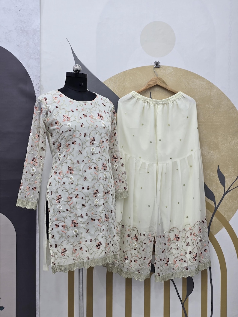 May include: A cream-colored embroidered tunic and matching palazzo pants set. The tunic features long sleeves and a floral pattern with pink accents. The palazzo pants have a similar embroidered border. The outfit is displayed on a mannequin.