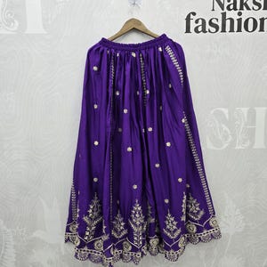 Peut inclure: Une jupe violette vibrante avec des broderies dor&eacute;es complexes. La jupe est suspendue &agrave; un cintre en bois. La broderie pr&eacute;sente des motifs floraux et g&eacute;om&eacute;triques sur l'ourlet et des lignes verticales. Le tissu de la jupe a un l&eacute;ger &eacute;clat.