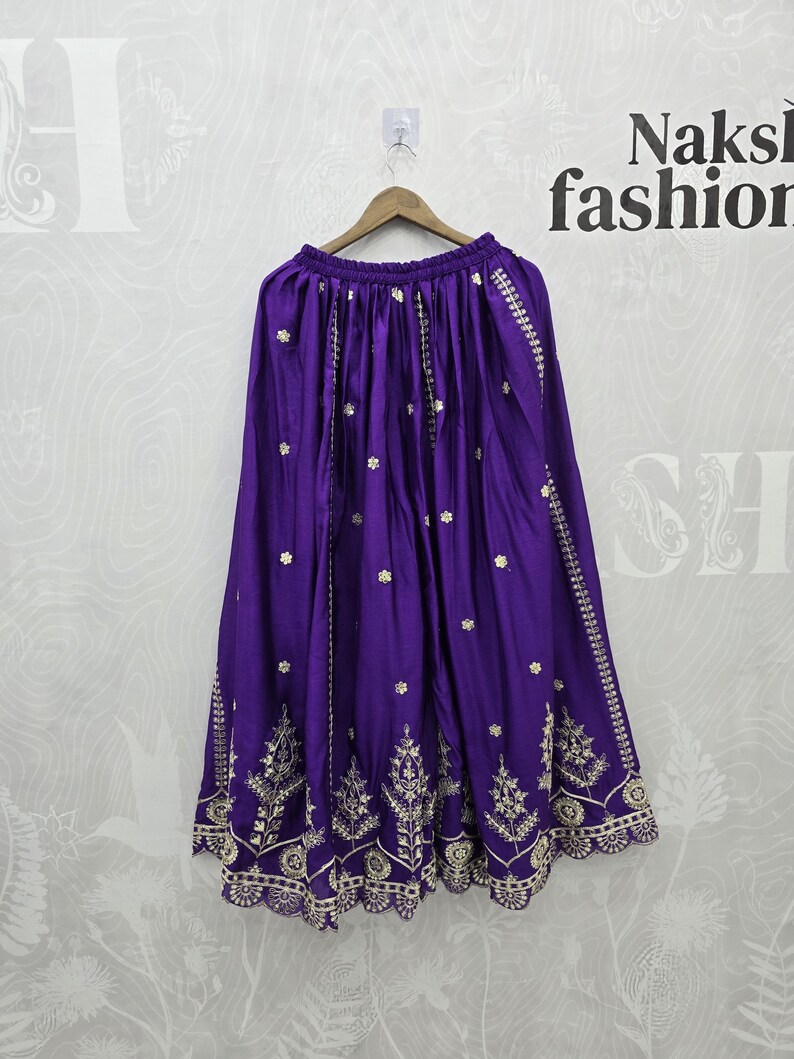 Peut inclure: Une jupe violette vibrante avec des broderies dor&eacute;es complexes. La jupe est suspendue &agrave; un cintre en bois. La broderie pr&eacute;sente des motifs floraux et g&eacute;om&eacute;triques sur l'ourlet et des lignes verticales. Le tissu de la jupe a un l&eacute;ger &eacute;clat.
