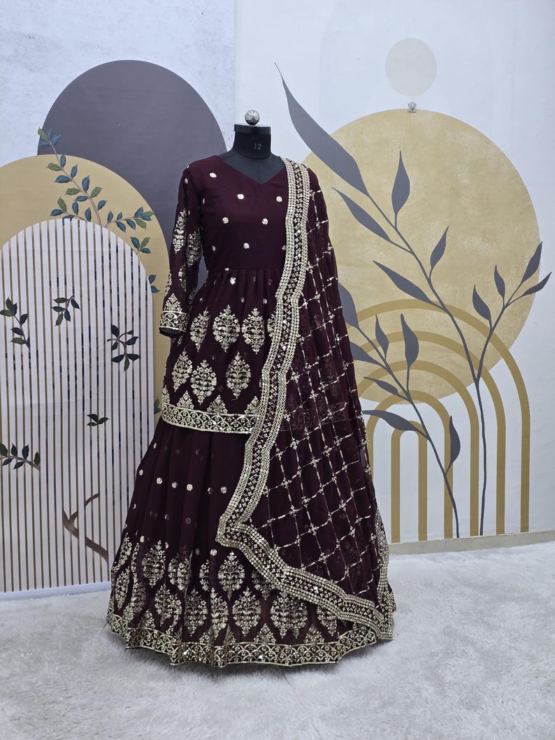 May include: A maroon-colored dress with gold embroidery and sequins. The dress has a long skirt and a fitted bodice. The dress is on a mannequin and has a matching dupatta.