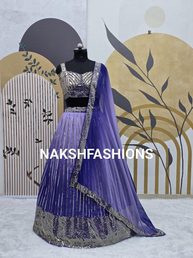 Op de afbeelding: Een paarse en zilveren, met pailletten bezette lehenga met een bijpassend crop top en dupatta. De lehenga heeft een verloopdesign, met een lichtere paarse kleur aan de bovenkant en een donkerdere paarse kleur aan de onderkant. De dupatta is doorschijnend en heeft een zilveren rand.