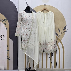 May include: A cream-colored, embroidered tunic and matching palazzo pants set. The tunic features long sleeves and a sheer overlay with floral embroidery. The palazzo pants are adorned with similar embroidery along the hemline. The outfit is displayed on a mannequin.