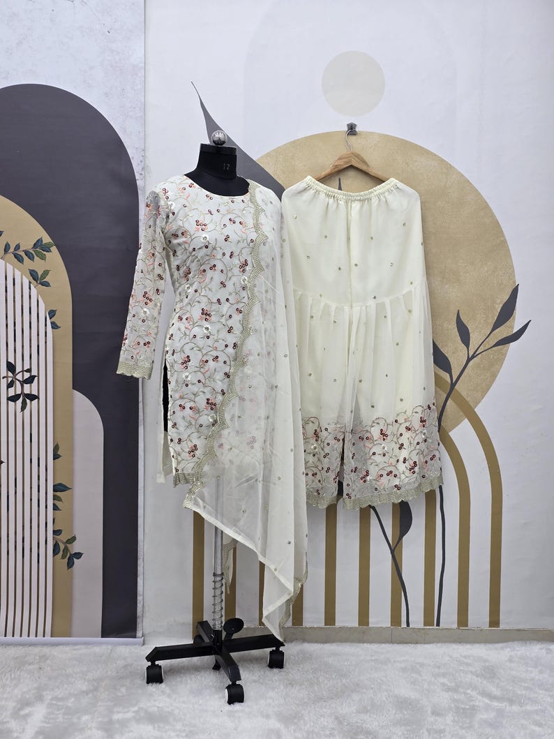 May include: A cream-colored, embroidered tunic and matching palazzo pants set. The tunic features long sleeves and a sheer overlay with floral embroidery. The palazzo pants are adorned with similar embroidery along the hemline. The outfit is displayed on a mannequin.
