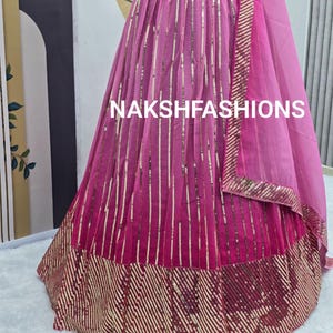 Op de afbeelding: Een roze en gouden paillettenrok met een gelaagd ontwerp. De rok heeft een zwarte tailleband en een doorschijnende roze dupatta met gouden bies. De rok is versierd met verticale rijen pailletten en een horizontale band van pailletten aan de zoom. De tekst "NAKSHFASHIONS" is zichtbaar op de rok.