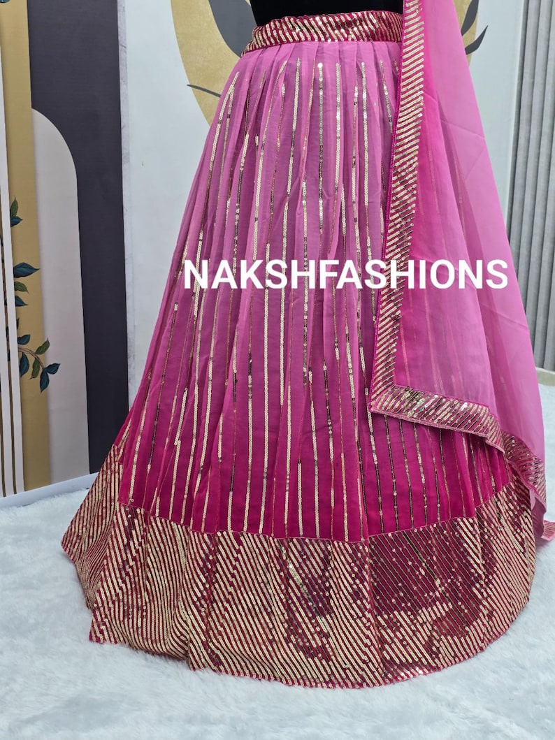 Op de afbeelding: Een roze en gouden paillettenrok met een gelaagd ontwerp. De rok heeft een zwarte tailleband en een doorschijnende roze dupatta met gouden bies. De rok is versierd met verticale rijen pailletten en een horizontale band van pailletten aan de zoom. De tekst "NAKSHFASHIONS" is zichtbaar op de rok.