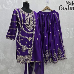 Peut inclure: Un ensemble violet vif comprenant une tunique, une jupe et une &eacute;charpe. La tunique et la jupe sont orn&eacute;es de broderies dor&eacute;es complexes. La tenue est pr&eacute;sent&eacute;e sur un mannequin et un cintre, mettant en valeur le savoir-faire et le design.
