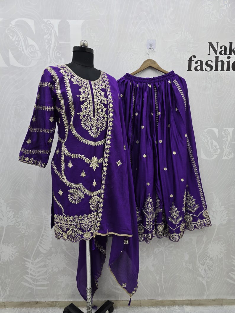 Peut inclure: Un ensemble violet vif comprenant une tunique, une jupe et une &eacute;charpe. La tunique et la jupe sont orn&eacute;es de broderies dor&eacute;es complexes. La tenue est pr&eacute;sent&eacute;e sur un mannequin et un cintre, mettant en valeur le savoir-faire et le design.