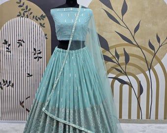 May include: A light blue embroidered lehenga with a matching crop top. The skirt is made of a sheer fabric with silver embroidery and sequins. The top is also embroidered with silver sequins. The lehenga is worn on a mannequin against a white and gold background.
