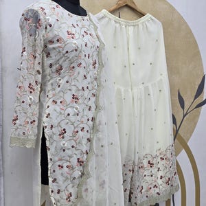 May include: A white embroidered outfit, including a long-sleeved top and wide-leg pants, displayed on a mannequin. The outfit features floral embroidery in shades of red and pink, with a scalloped trim. The pants have scattered embellishments.