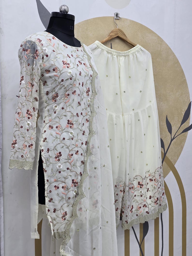 May include: A white embroidered outfit, including a long-sleeved top and wide-leg pants, displayed on a mannequin. The outfit features floral embroidery in shades of red and pink, with a scalloped trim. The pants have scattered embellishments.