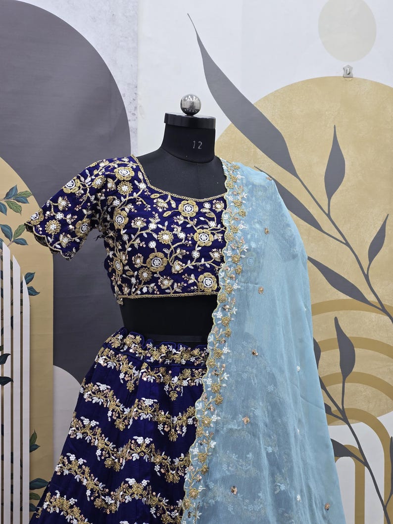 May include: A blue and gold embroidered lehenga with a matching blue and gold embroidered choli. The lehenga has a light blue sheer dupatta with gold embroidery.