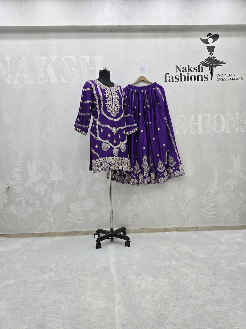 Peut inclure: Une tenue violette brod&eacute;e, compos&eacute;e d'un haut &agrave; manches longues et d'une jupe, pr&eacute;sent&eacute;e sur un mannequin et un cintre. La tenue pr&eacute;sente des broderies dor&eacute;es complexes. La marque "Naksh fashions" est visible en arri&egrave;re-plan.