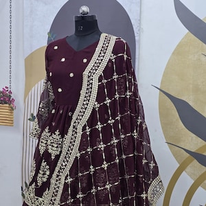 May include: A maroon-colored dress with gold embroidery and sequins. The dress has a V-neckline and three-quarter sleeves. It is paired with a matching maroon dupatta with gold embroidery and sequins.
