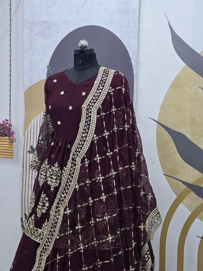 May include: A maroon-colored dress with gold embroidery and sequins. The dress has a V-neckline and three-quarter sleeves. It is paired with a matching maroon dupatta with gold embroidery and sequins.