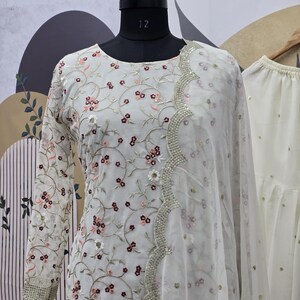 May include: A white embroidered outfit with a floral pattern. The long-sleeved top features delicate embroidery in shades of pink and burgundy. A sheer scarf with a scalloped edge and silver embellishments drapes over the outfit. The outfit is displayed on a mannequin.