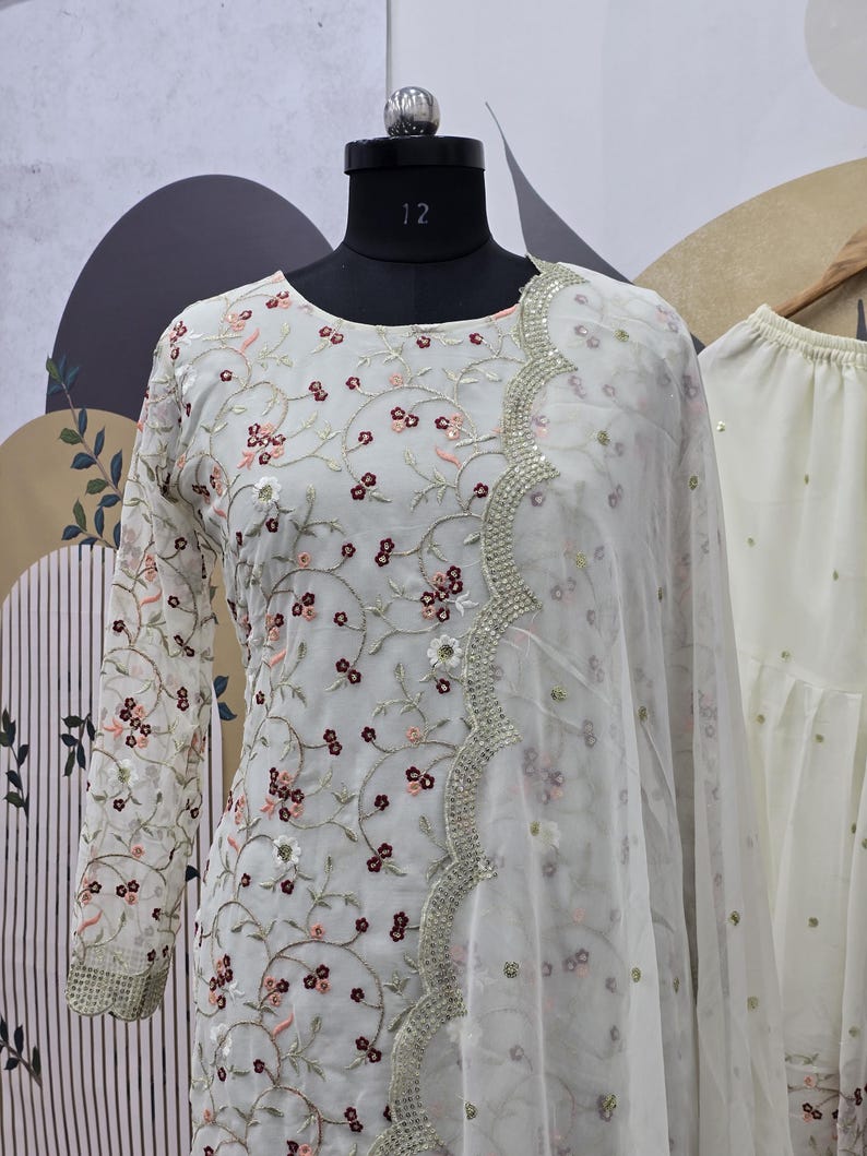 May include: A white embroidered outfit with a floral pattern. The long-sleeved top features delicate embroidery in shades of pink and burgundy. A sheer scarf with a scalloped edge and silver embellishments drapes over the outfit. The outfit is displayed on a mannequin.