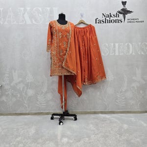 May include: An orange embroidered salwar kameez set displayed on a mannequin and hanger. The set includes a long-sleeved tunic, a matching skirt, and a scarf. The outfit is embellished with gold embroidery. The brand logo "Naksh fashions" is visible.