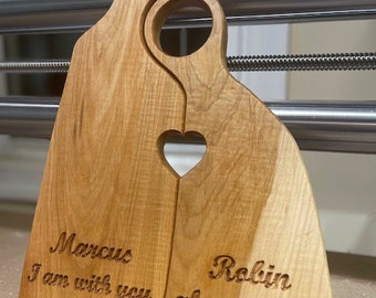 May include: Two wooden cutting boards interlock to form a heart shape. The boards are light brown with a natural wood grain. One board has the name "Marcus" and the phrase "I am with you" engraved. The other board has the name "Robin", the word "always", and "Matt 28:20" engraved.