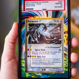 K&ouml;nnte beinhalten: Eine bewertete 2005 Pokemon EX Deoxys Rayquaza Gold Star Karte. Die Karte befindet sich in einer Schutzh&uuml;lle mit einem PSA-Etikett. Die Karte zeigt eine schwarz-gr&uuml;ne Illustration von Rayquaza mit dem Text "Rayquaza" und "90 HP".