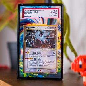 K&ouml;nnte beinhalten: Eine gerahmte Pok&eacute;mon-Karte mit Rayquaza, eine holografische Gold Star-Karte aus dem 2005 EX Deoxys-Set. Die Karte ist von PSA mit GEM MT 10 bewertet. Das Design der Karte zeigt ein schwarzes und rotes drachenartiges Wesen mit dem Text "Rayquaza" und "90 HP".