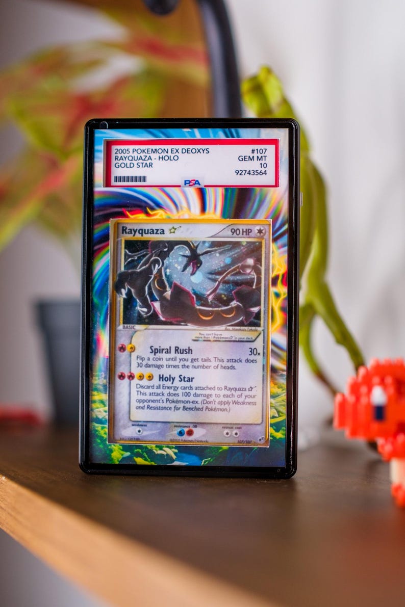 K&ouml;nnte beinhalten: Eine gerahmte Pok&eacute;mon-Karte mit Rayquaza, eine holografische Gold Star-Karte aus dem 2005 EX Deoxys-Set. Die Karte ist von PSA mit GEM MT 10 bewertet. Das Design der Karte zeigt ein schwarzes und rotes drachenartiges Wesen mit dem Text "Rayquaza" und "90 HP".