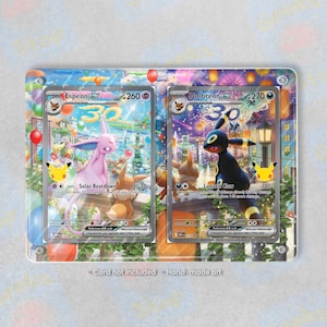 May include: A rectangular display frame containing two trading cards featuring Espeon ex and Umbreon ex. The cards show colourful illustrations of the creatures in a festive setting with fireworks and balloons. Text at the bottom reads Card not included.