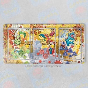 May include: A rectangular acrylic display frame featuring three Pokémon trading card illustrations of Chikorita, Cyndaquil, and Totodile set against a yellow autumn leaf background. The frame has silver corner screws and text reading Cards not included - Hand-made art.