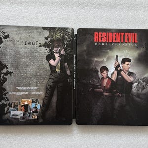 May include: A Resident Evil Code: Veronica video game case, open to display the front and back covers. The front cover features the game title in red, with characters holding guns. The back cover has text and images.