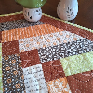 May include: A quilted table runner with a patchwork design in shades of brown, orange, and green. Two ceramic mushroom figurines with white stems and colorful caps sit on the runner. The runner features text and floral patterns.