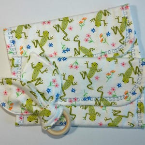 May include: Two white fabric burp cloths with a green frog and flower pattern. The cloths are folded and stacked on top of each other. A wooden teething ring is in front of the cloths.